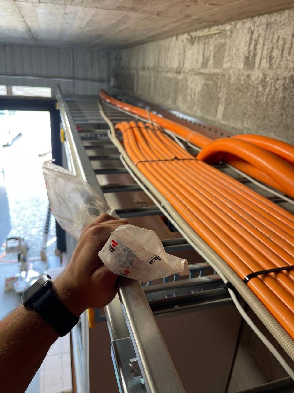 Une main tenant un sac près d'une échelle supportant des câbles orange dans un bâtiment en béton.
