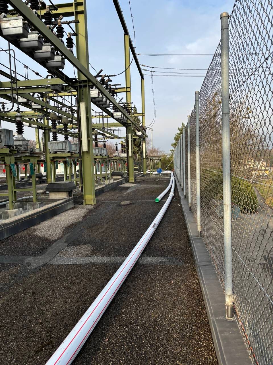Poste de transformation électrique avec lignes aériennes, structures métalliques et clôture. Un tuyau blanc longe la clôture.