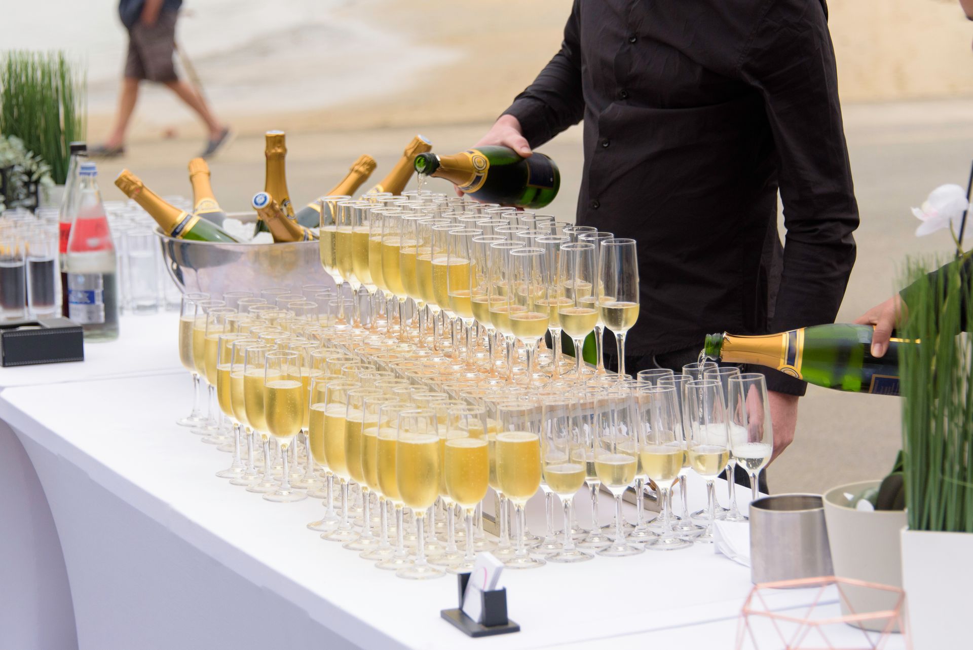 Des bouteilles de champagne sont versées dans des rangées de flûtes à champagne sur une table blanche à l'extérieur.