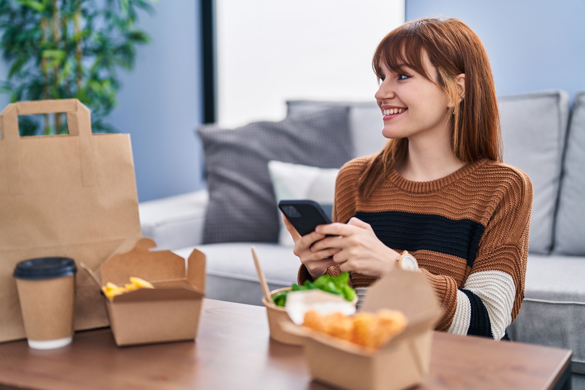 Une femme utilise son téléphone à une table où sont posés des plats à emporter et du café, dans un salon.