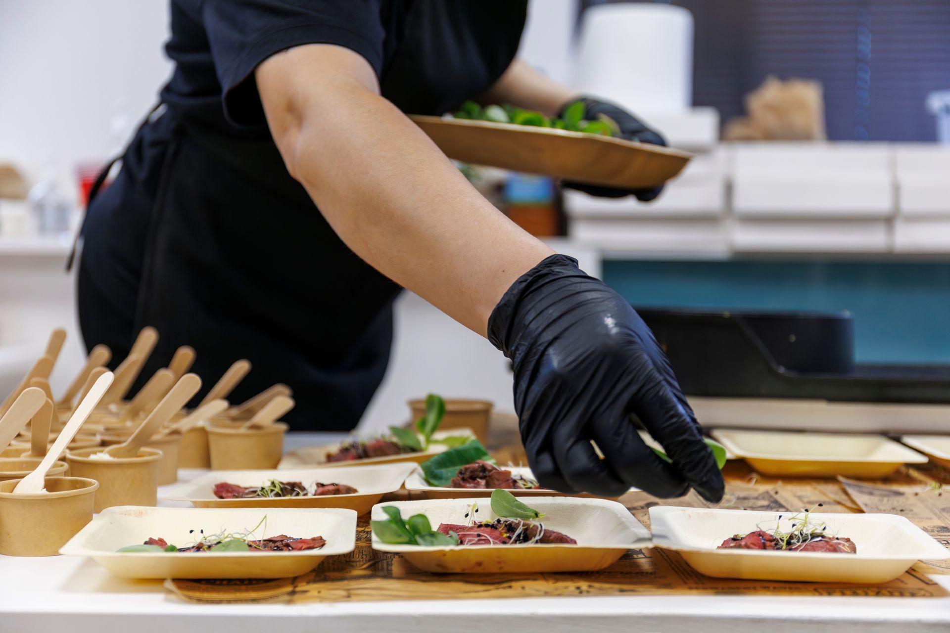 Un employé de cuisine dispose de petites entrées sur des plateaux en bois dans une cuisine, portant des gants noirs.