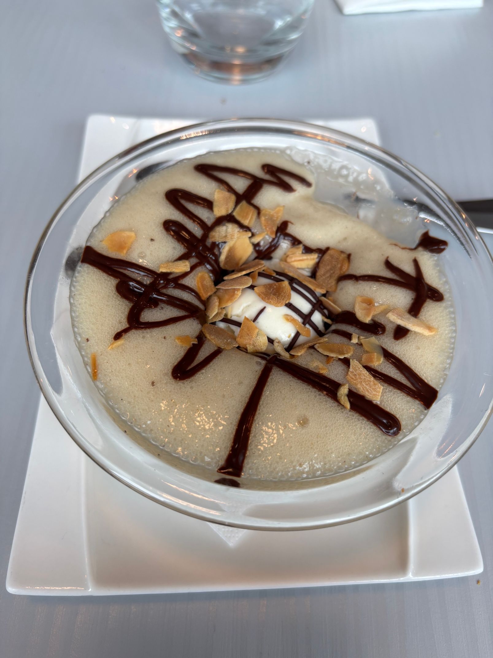 Dessert crémeux servi dans un bol en verre, garni de tranches de banane et d'un filet de chocolat, sur une assiette blanche.