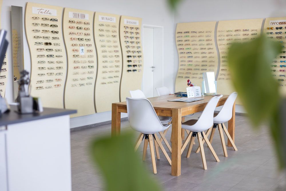 Eine Optikerpraxis: Brillenaufstellung an der Wand, ein Holztisch mit weißen Stühlen und eine Pflanze im Vordergrund.