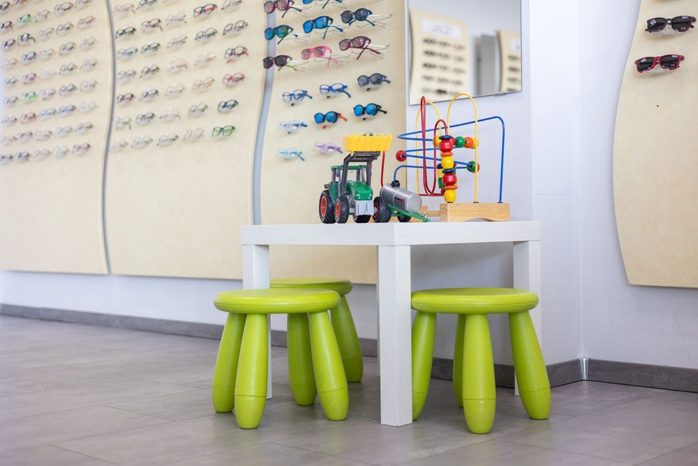 Ein Kinderspielbereich in einer Optikerpraxis mit einem Tisch, Hockern, Spielzeug und ausgestellten Brillen.