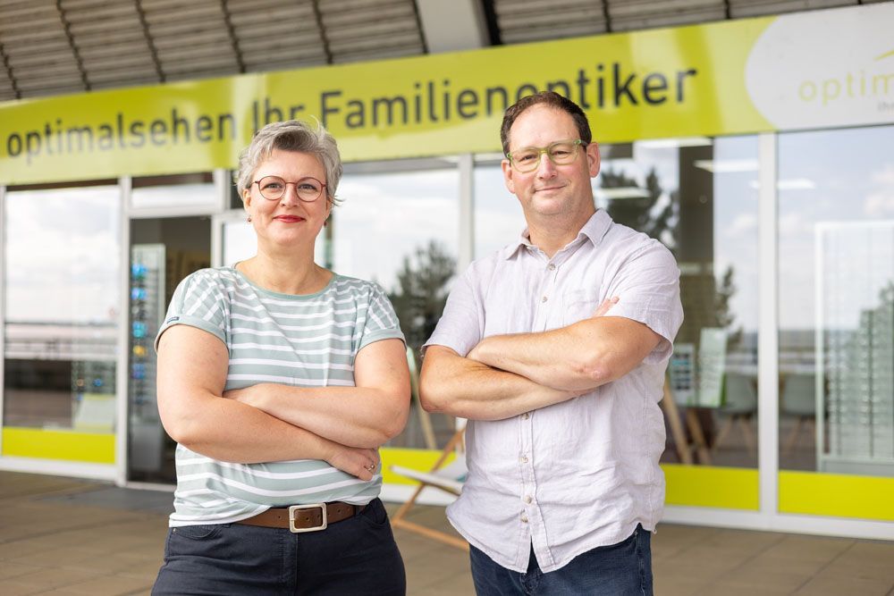 Zwei Personen, eine Frau und ein Mann, stehen mit verschränkten Armen vor einem Optikergeschäft.
