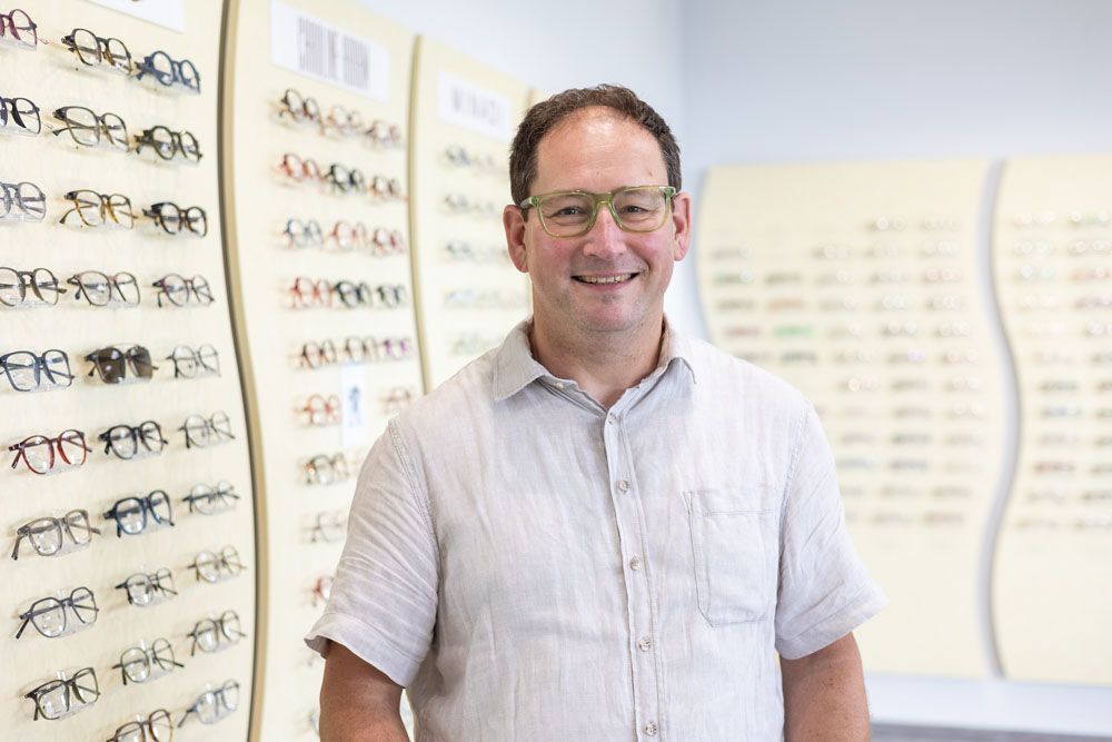 Mann mit Brille lächelt in einem Optikergeschäft, umgeben von Brillengestellen.