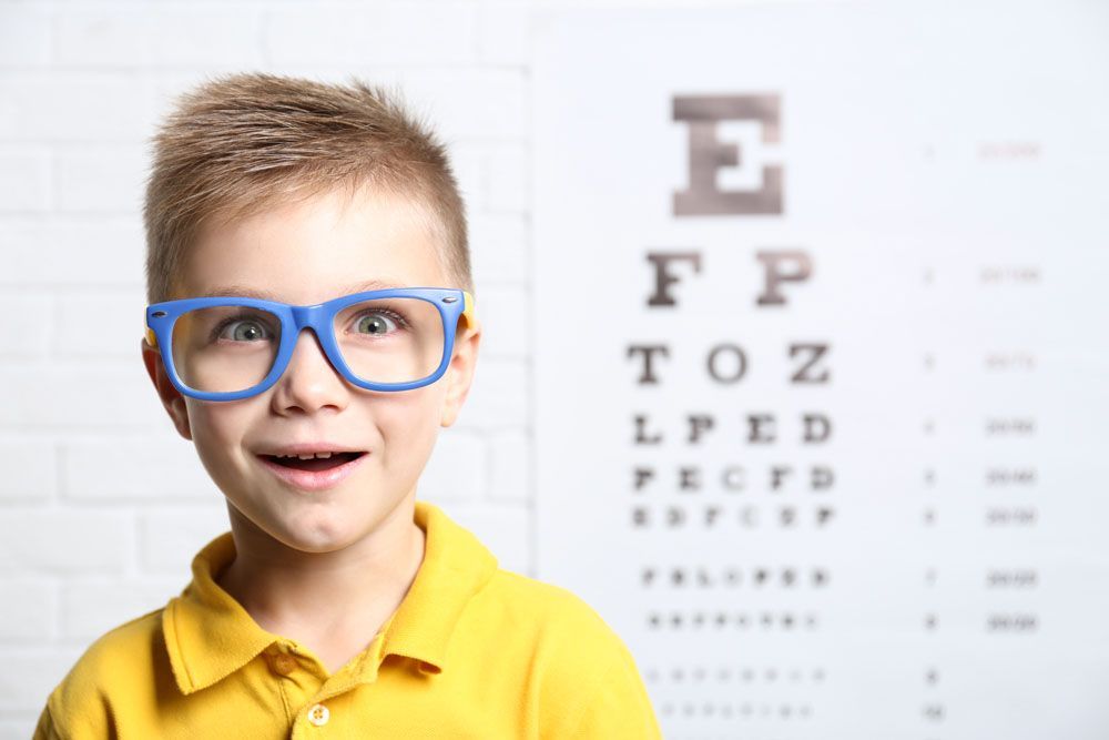 Junge mit blauer Brille lächelt vor einer Sehtafel.