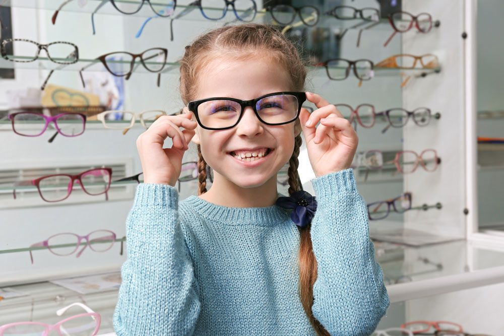 Mädchen mit Zöpfen lächelt, während sie in einem Optikergeschäft eine schwarze Brille anprobiert.