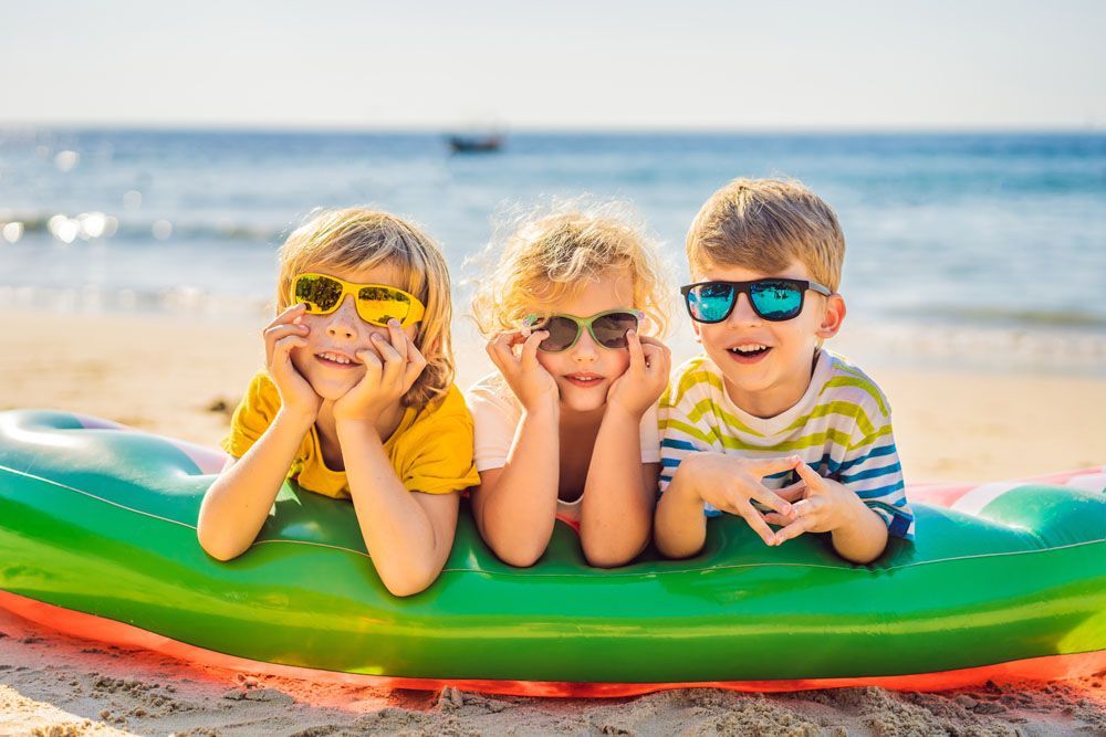 Drei Kinder mit Sonnenbrillen liegen lächelnd auf einem Schwimmreifen am Strand.