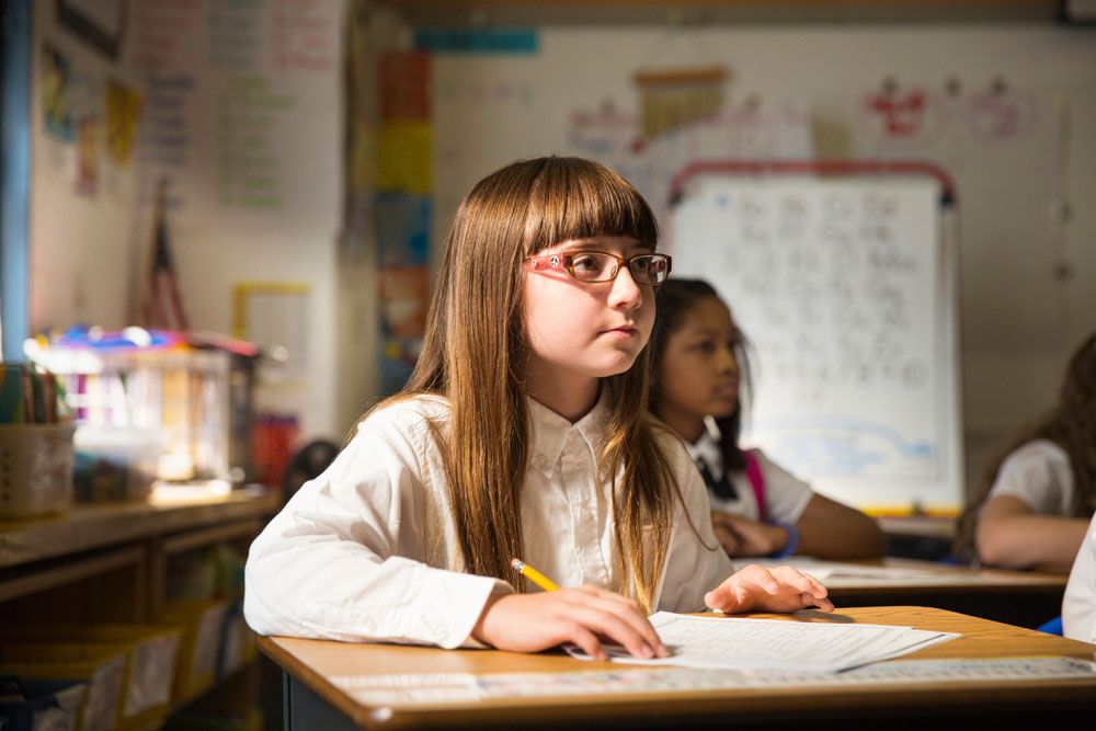 Mädchen mit Brille schreibt am Schreibtisch im Klassenzimmer, andere Schüler im Hintergrund.