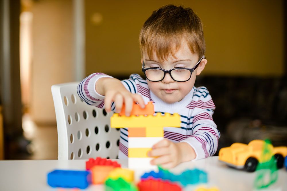 Ein kleiner Junge mit Down-Syndrom und Brille baut an einem Tisch mit bunten Bauklötzen.