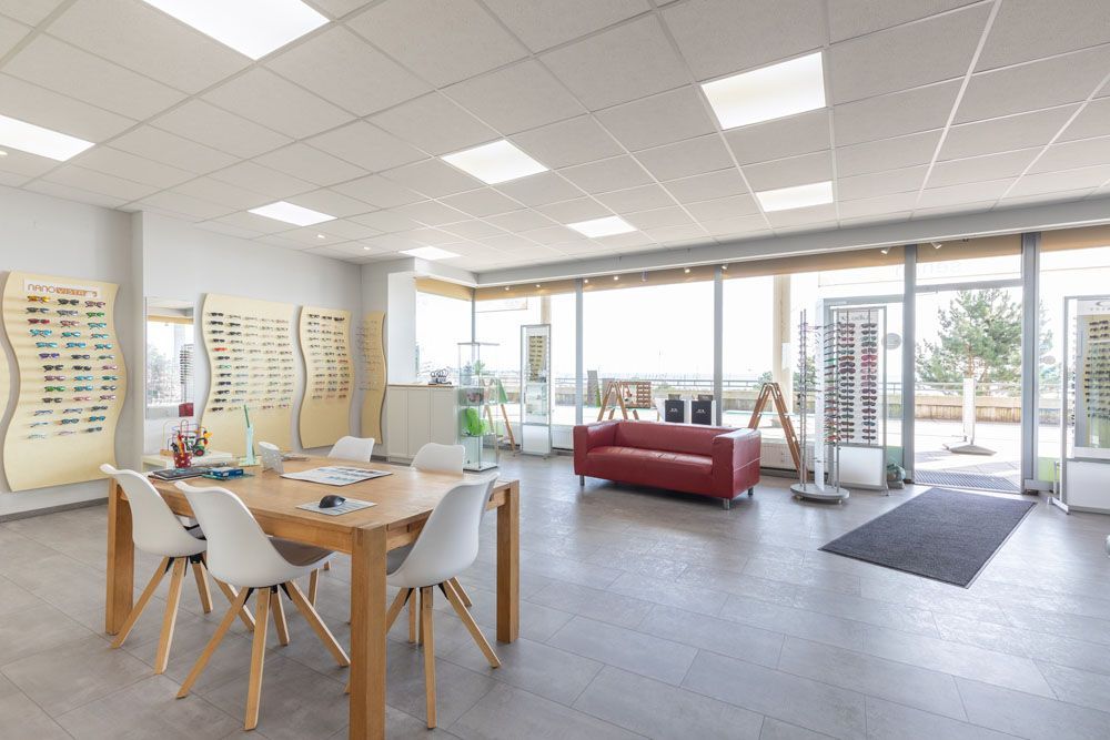 Modernes Optikergeschäft mit Brillenauslagen, einem roten Sofa und einem Tisch mit Stühlen. Große Fenster sorgen für Tageslicht.