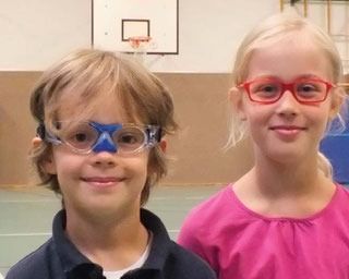 Zwei lächelnde Kinder, ein Junge mit einer Schutzbrille mit blauem Rahmen und ein Mädchen mit einer Brille mit rotem Rahmen. Drinnen, in der Nähe eines Basketballkorbs.