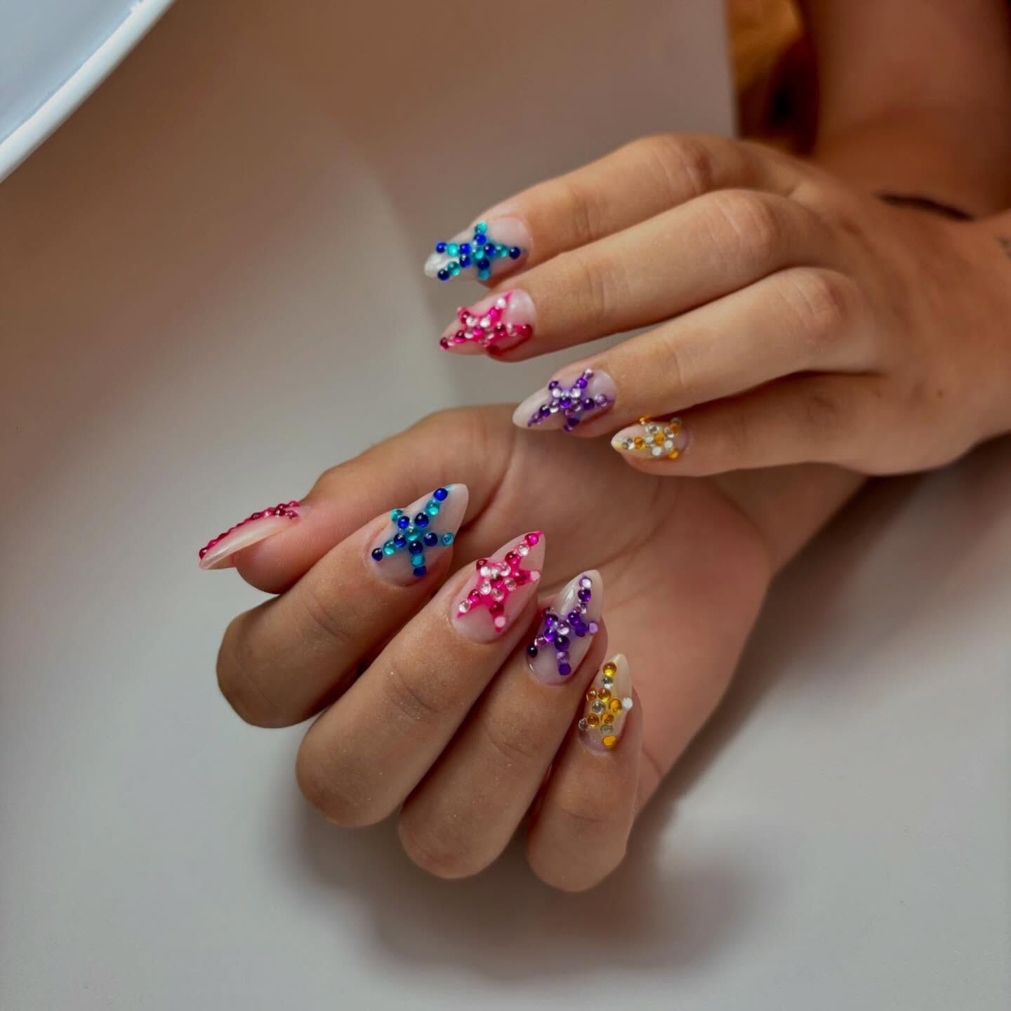 Manos con uñas almendradas decoradas con coloridos diseños de estrellas de pedrería sobre una base neutra, sostenidas sobre una superficie blanca.