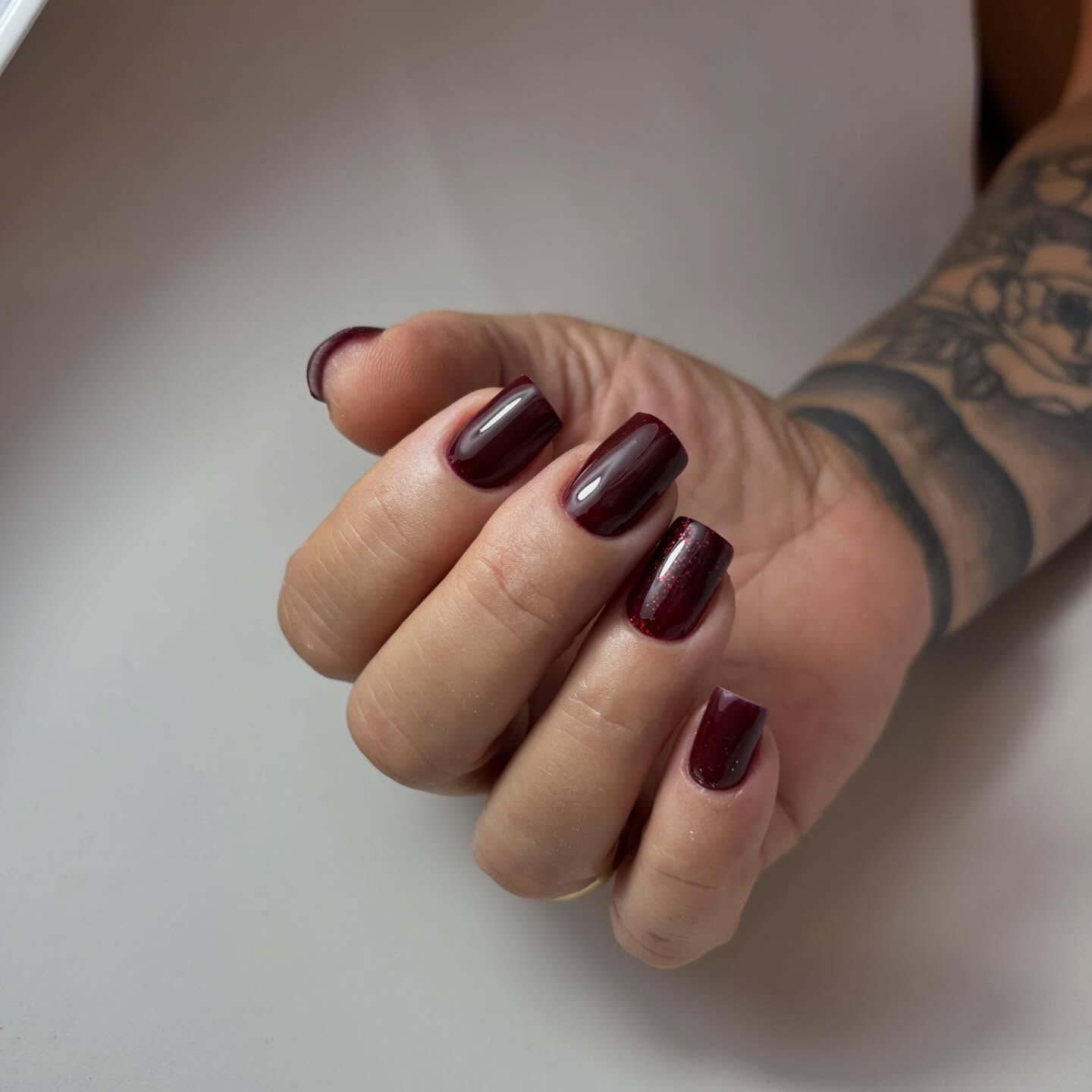 Una mano con uñas cuadradas pintadas con un esmalte brillante de color burdeos oscuro, y con parte de un tatuaje visible en el brazo.