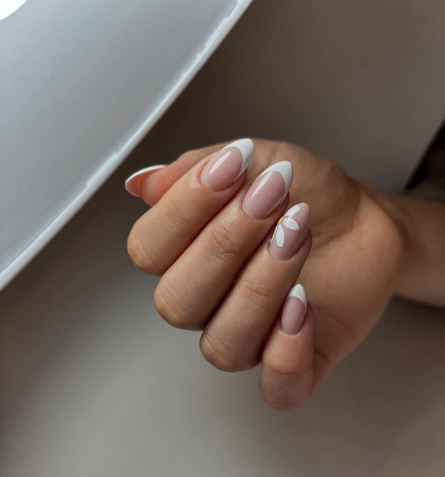 Una mano que luce una manicura en forma de almendra con una clásica punta francesa blanca y un pequeño adorno de margarita blanca en una de las uña.