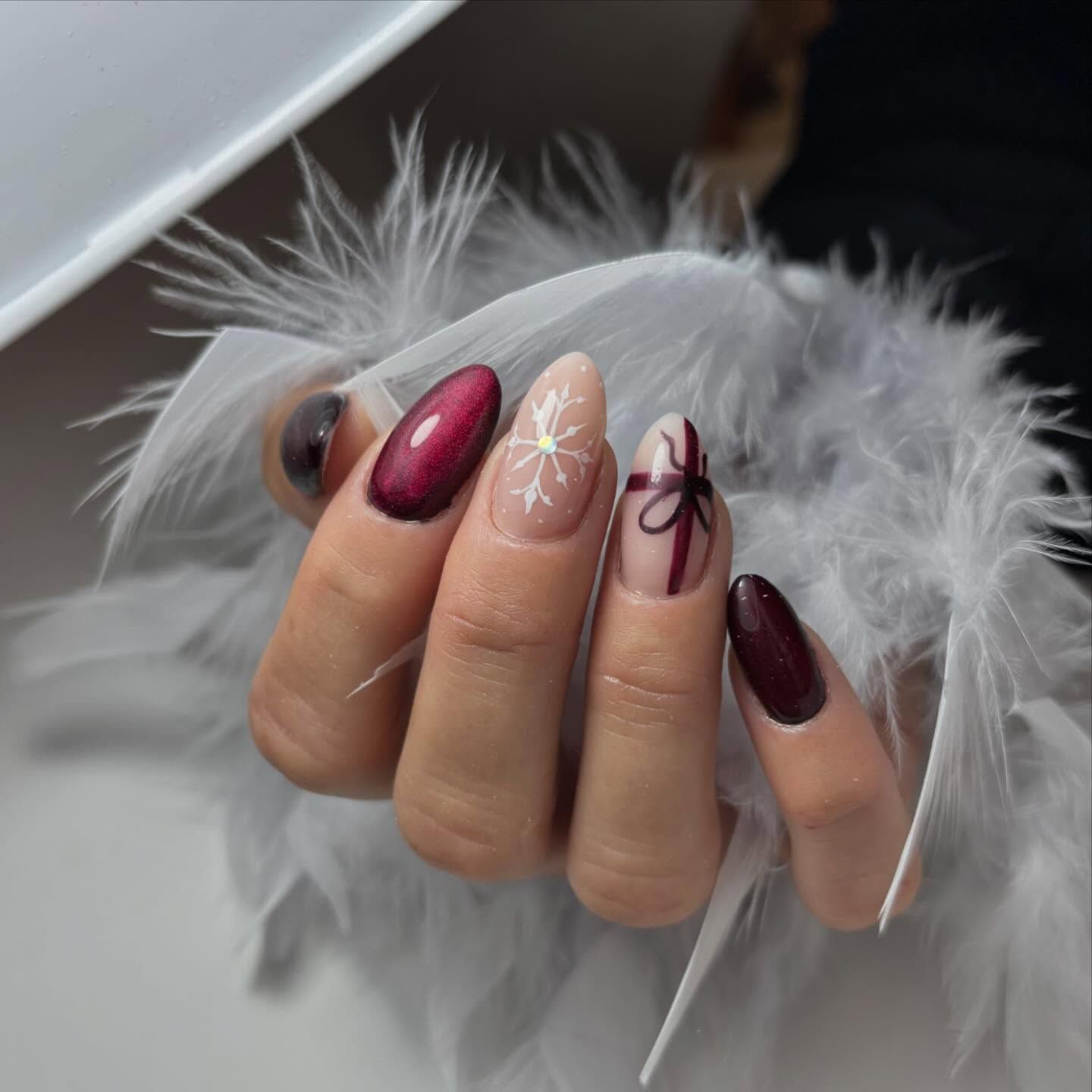 Manos con manicura impecable sobre un fondo de plumas blancas, con diseños de uñas ovaladas en rojo, copos de nieve y cintas de regalo.