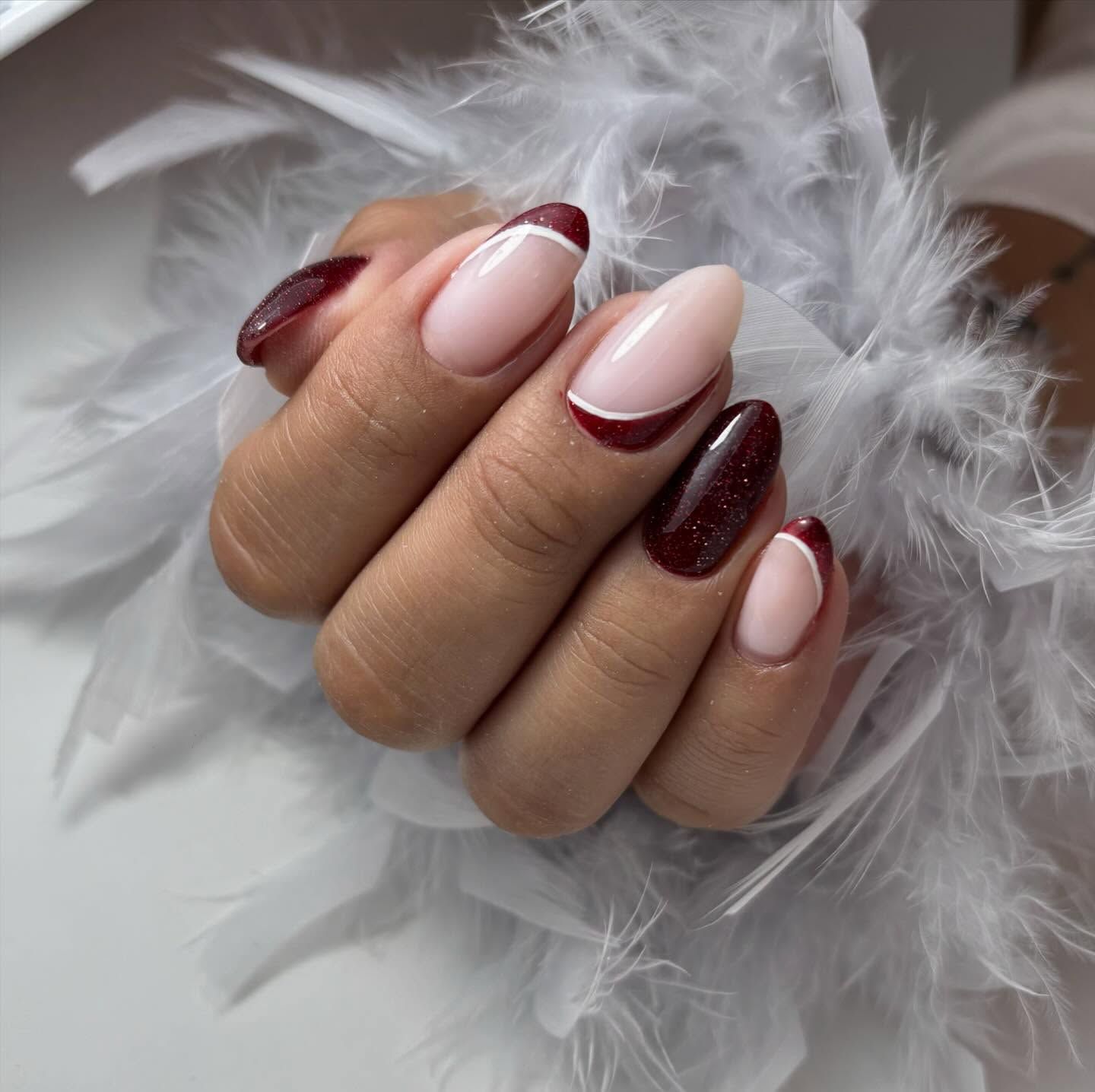 Una mano con uñas en forma de almendra que presentan alternancia de esmalte rojo oscuro con purpurina y una base color nude con una punta arqueada roja.