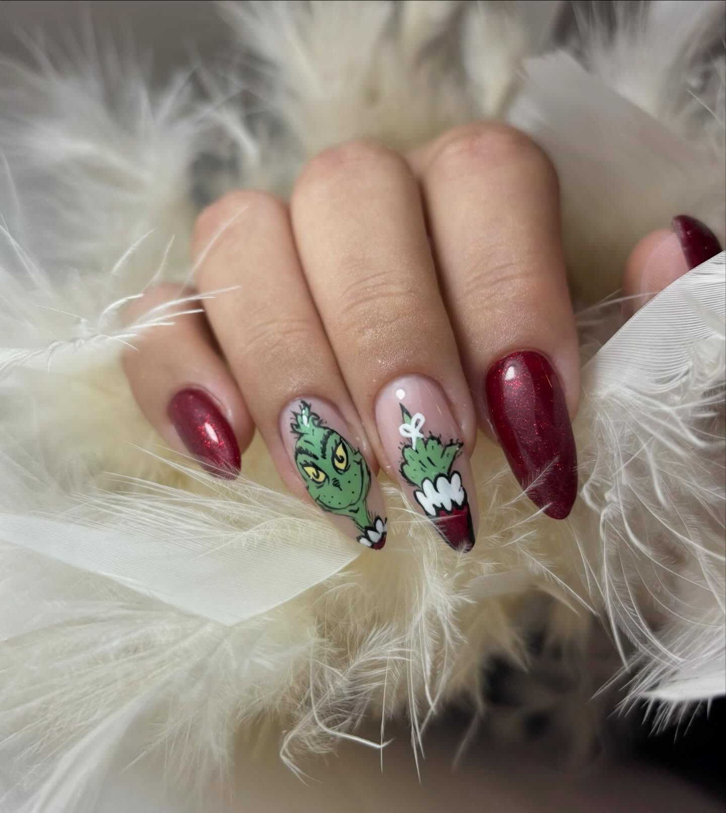 Uñas con forma de almendra, cuidadosamente arregladas, con dos uñas decoradas con motivos del Grinch, alternadas con esmalte rojo intenso brillante.