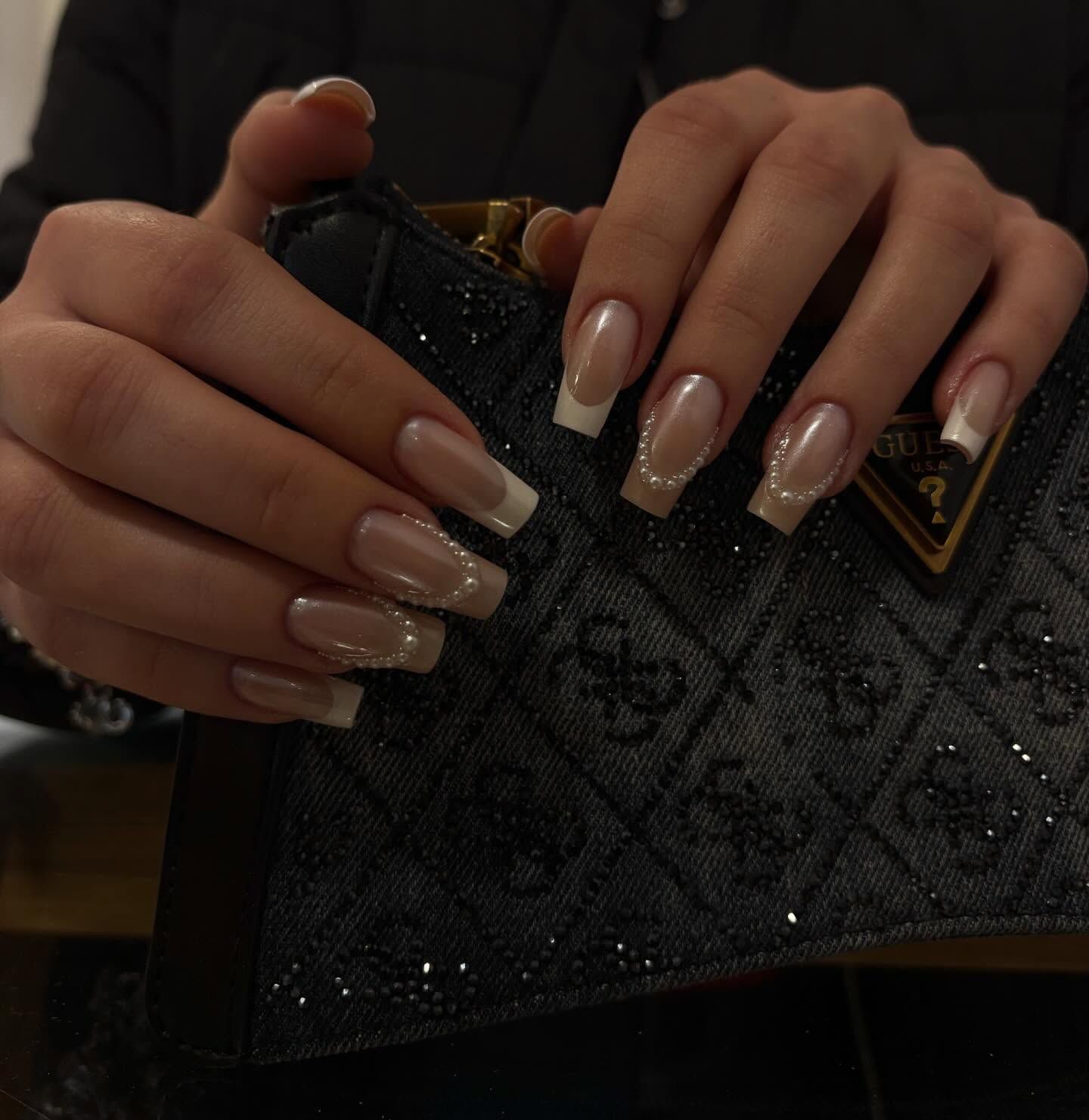 Manos con uñas largas y cuadradas con punta francesa y pequeños detalles de perlas, sosteniendo un bolso de mano gris oscuro con el monograma de Guess.