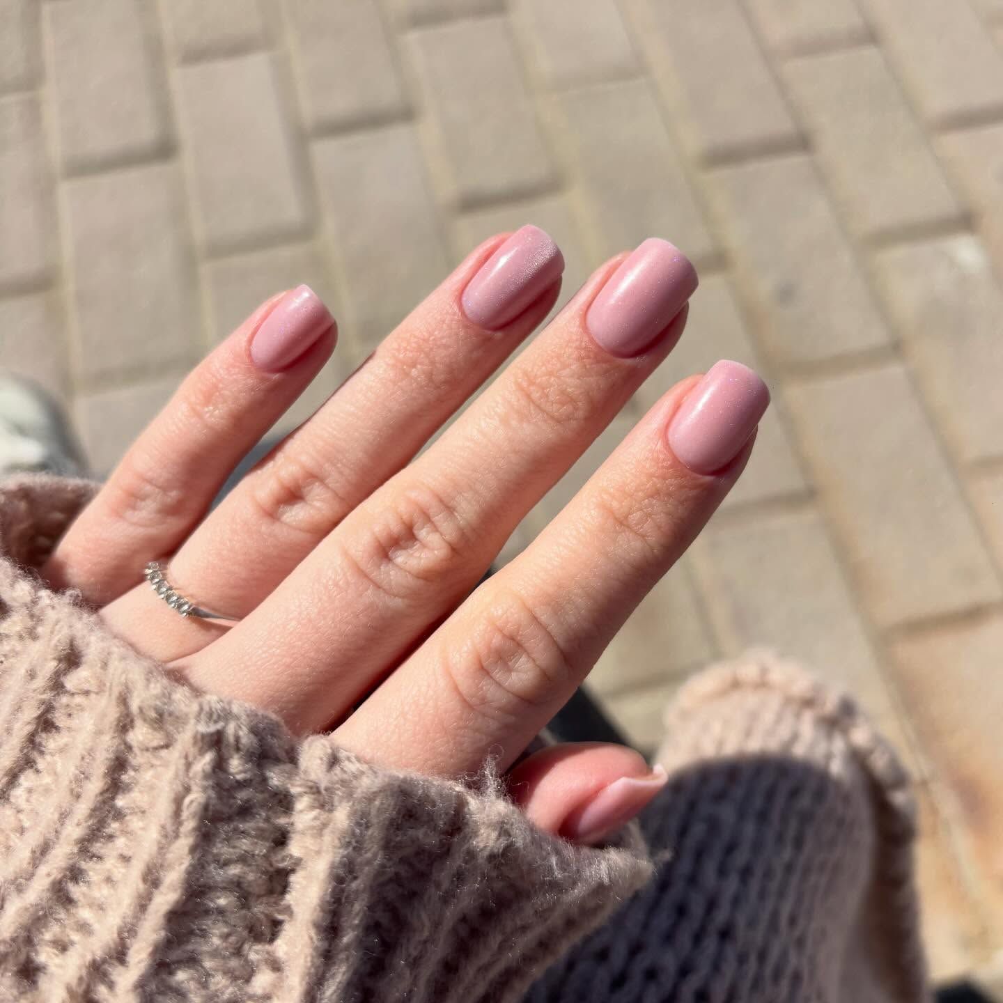 Una mano con un anillo de plata en el dedo, con las uñas pintadas de un rosa suave y corto, sobre un suéter texturizado de color marrón claro.