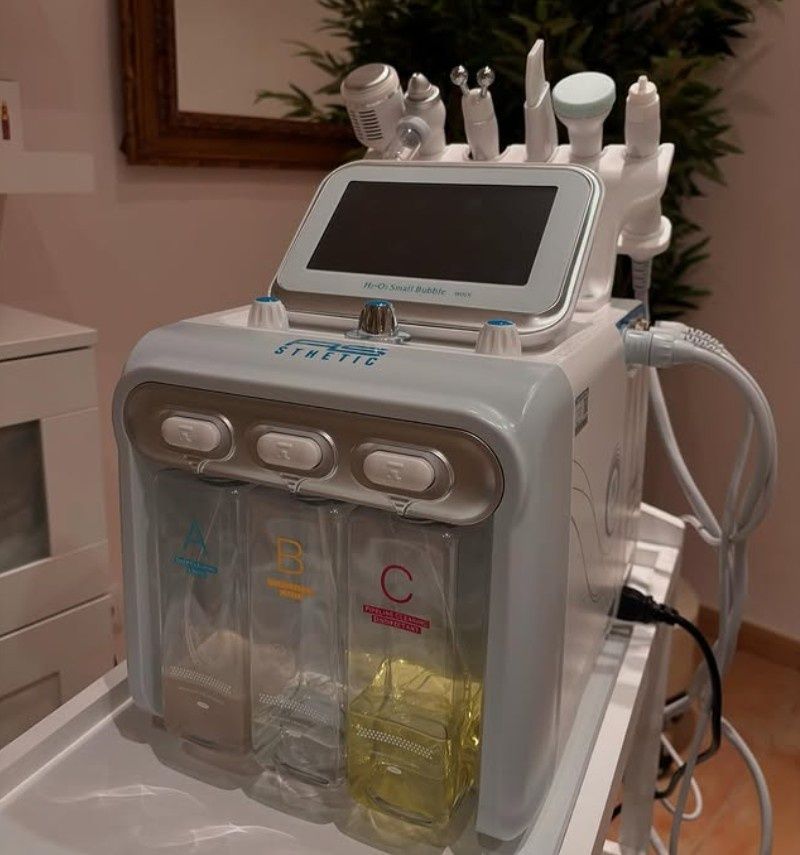 Un aparato facial estético multifuncional con pantalla táctil, varias puntas aplicadoras y tres depósitos de líquido etiquetados.