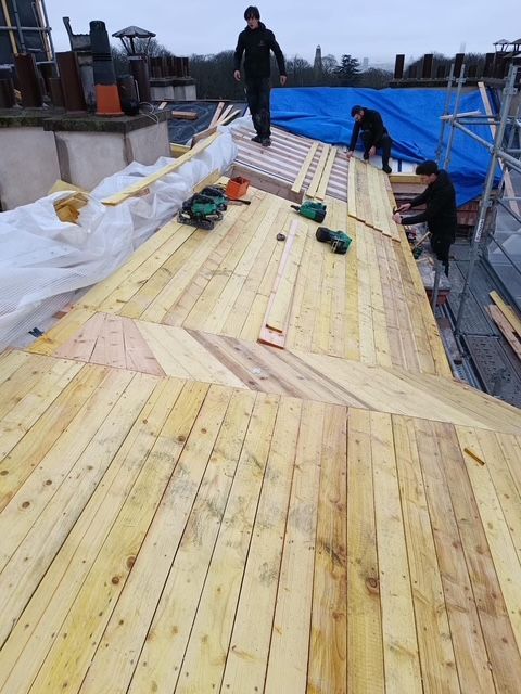 Des ouvriers installent des lames de bois sur une passerelle sur le toit, avec des outils et des bâches bleues le long des côtés.