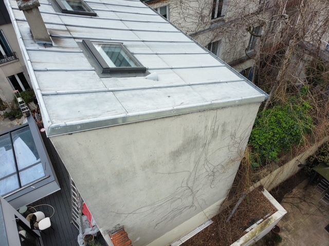 Vue aérienne du toit d'un bâtiment étroit, jouxtant un mur et une cour envahie par la végétation.