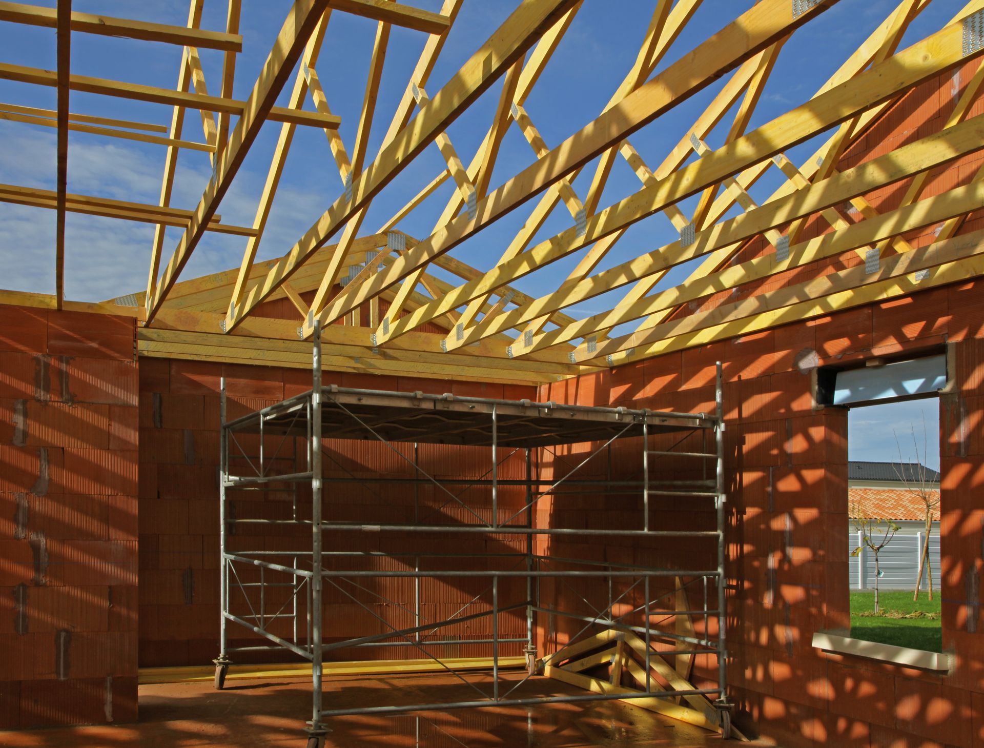 Intérieur d'une maison en briques en construction, avec échafaudages et poutres de toit apparentes, éclairé par la lumière du soleil.
