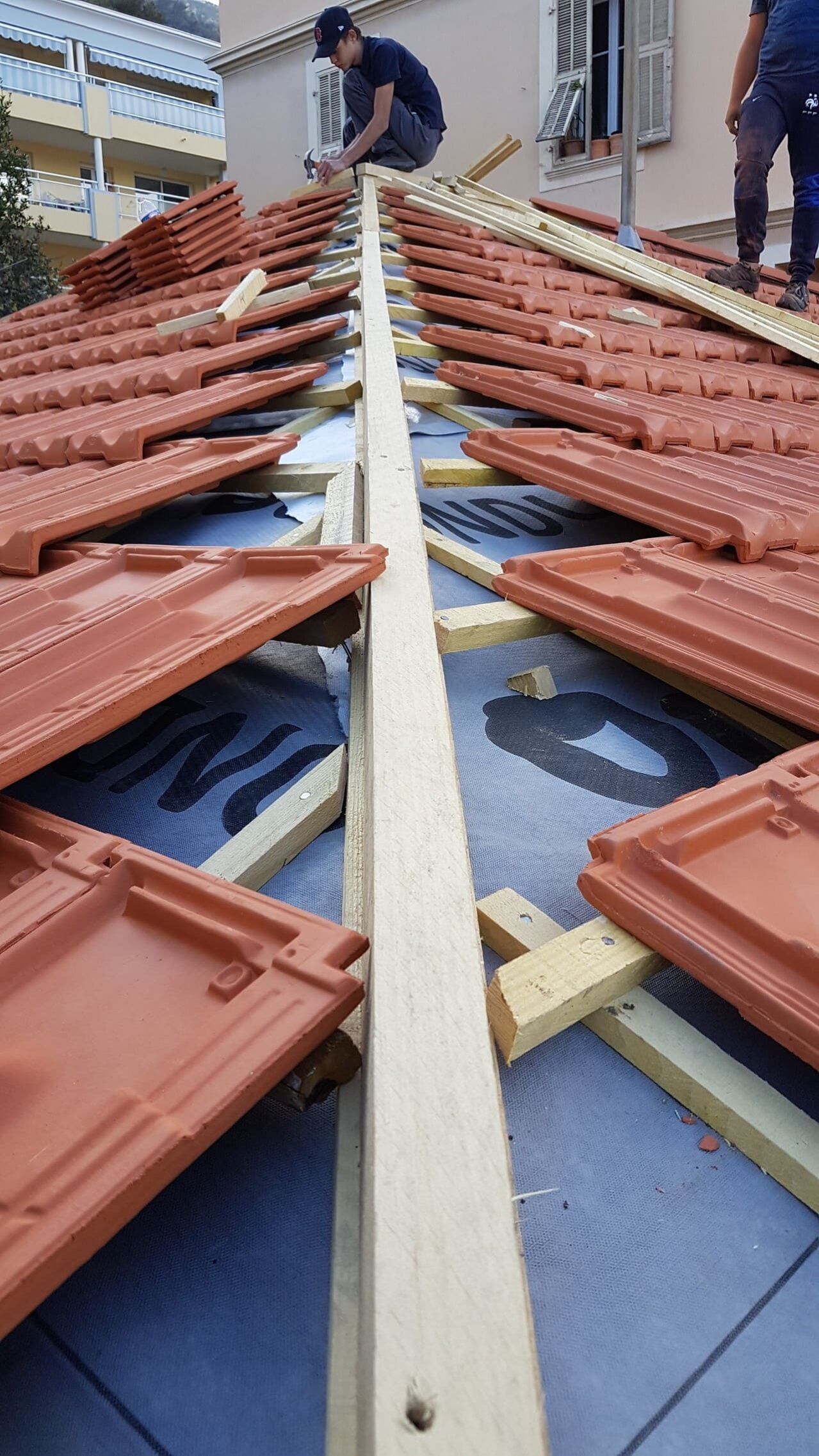 Des couvreurs posent des tuiles en terre cuite rouge sur un toit avec une poutre centrale en bois et une sous-couche bleue.