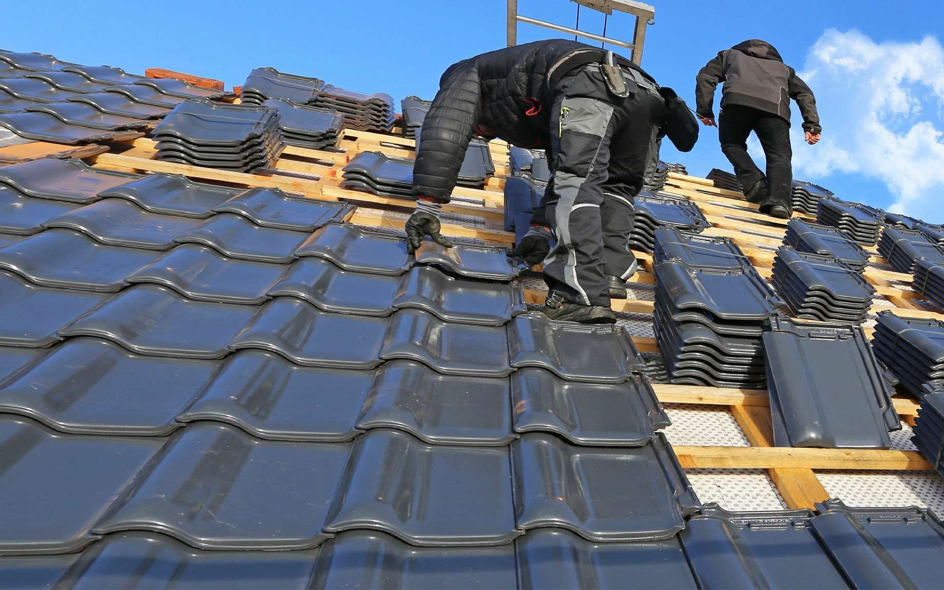 Des couvreurs installent des tuiles métalliques sombres sur un toit pentu sous un ciel bleu.