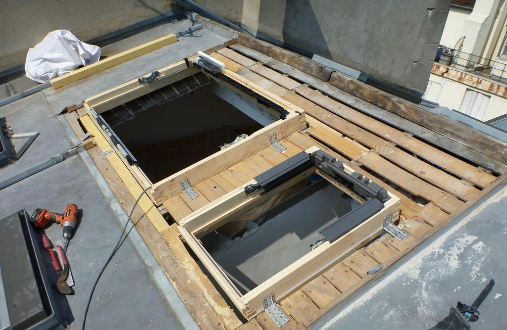 Chantier de construction sur un toit, avec une charpente en bois entourant de grandes ouvertures rectangulaires et des outils de chantier à proximité.
