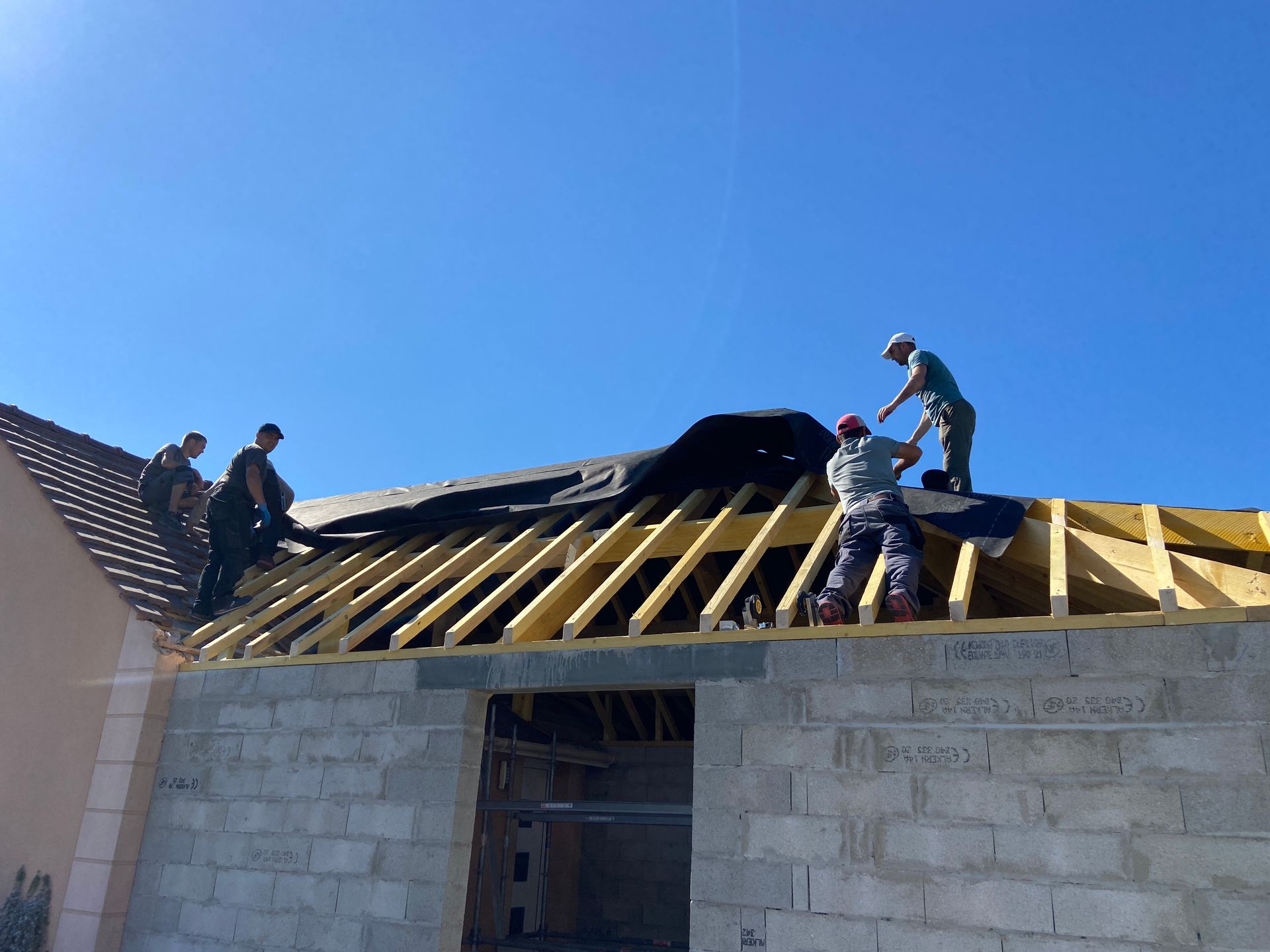 Des ouvriers construisent la charpente d'un toit sur un bâtiment en parpaings sous un ciel bleu dégagé.