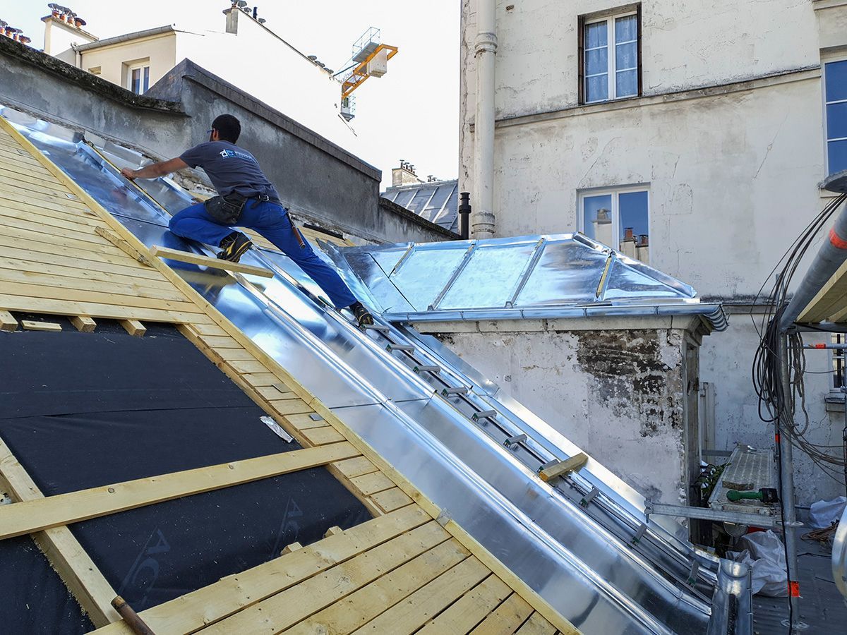 Un ouvrier installe des panneaux solaires sur un toit en pente, près d'une lucarne, dans une cour urbaine.