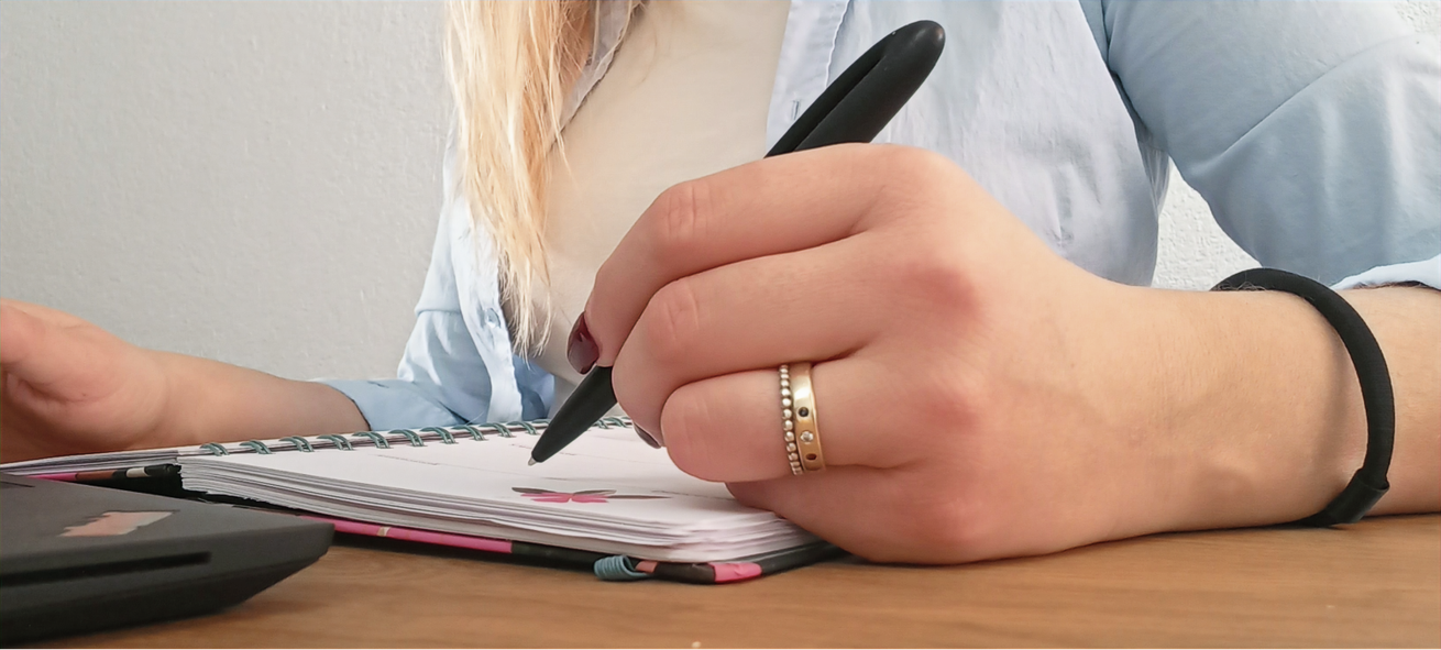 Handschriftliches Schreiben in einem Notizbuch am Schreibtisch, ein Taschenrechner in der Nähe und ein Stift in der Hand