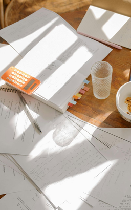 Papiere und Bücher auf einem Holztisch, Sonnenlicht, Taschenrechner, Glas, Snacks und Stift.