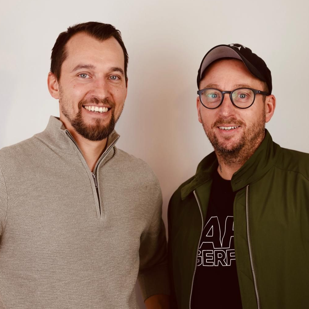 Zwei Männer stehen drinnen vor einer schlichten Wand, einer trägt einen grauen Pullover, der andere eine Jacke und Mütze.