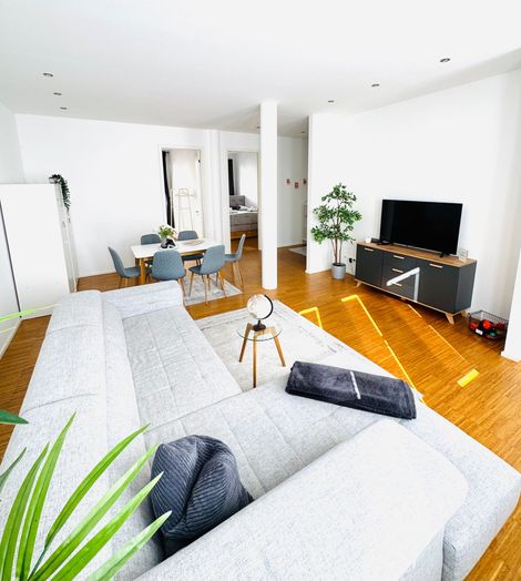 Helles, modernes Wohnzimmer mit weißem Sofa, Fernseher auf einem Ständer, Pflanzen und Holzboden, gesehen von hinter einem Sofa.