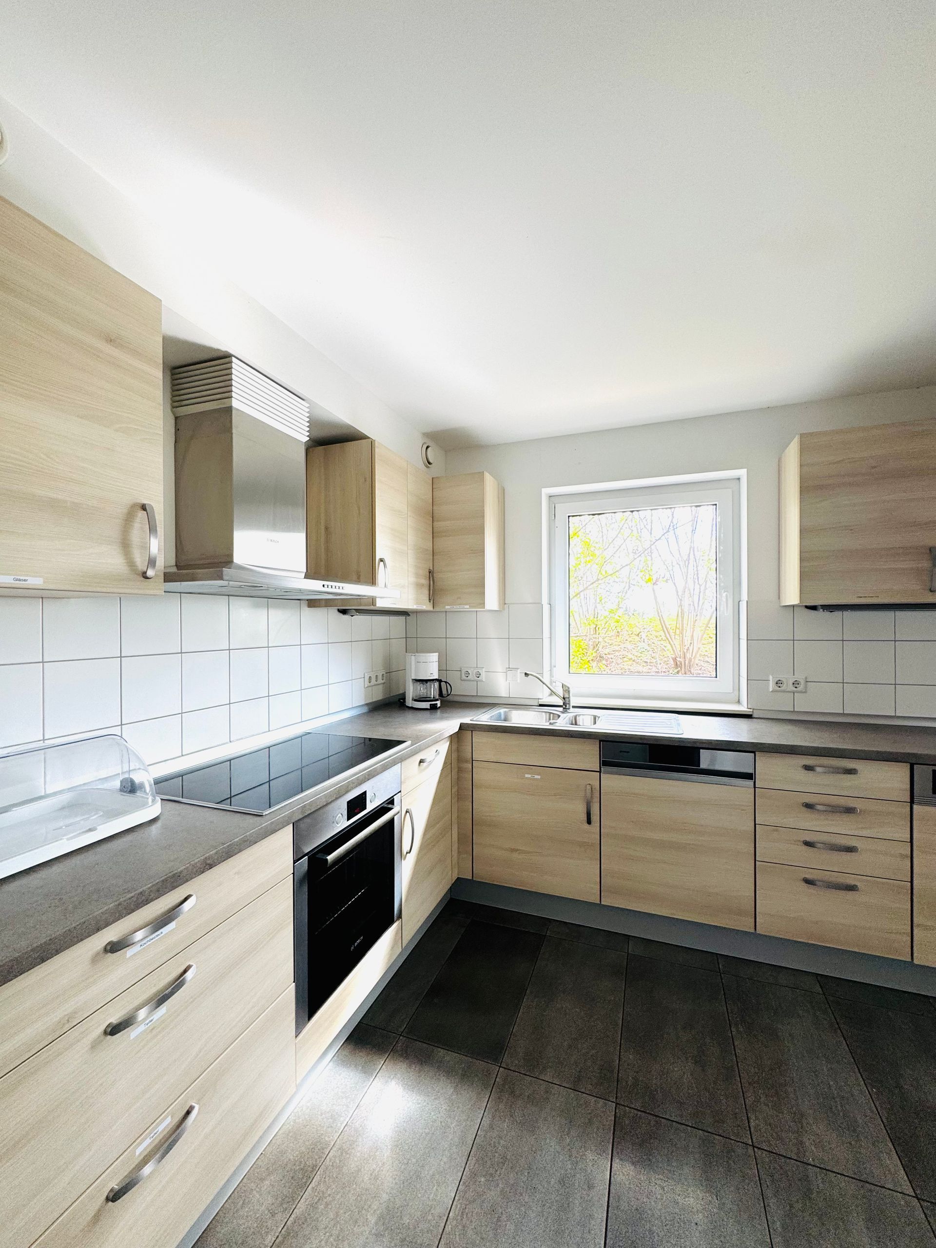 Moderne Küche mit hellen Holzschränken, weißem Fliesenspiegel, dunklem Boden und einem Fenster über der Spüle