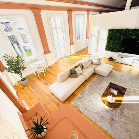 Helles Loft-Wohnzimmer mit weißer Eckcouch, Holzboden, großen Fenstern und einem Fernseher an einer grünen Wandverkleidung.