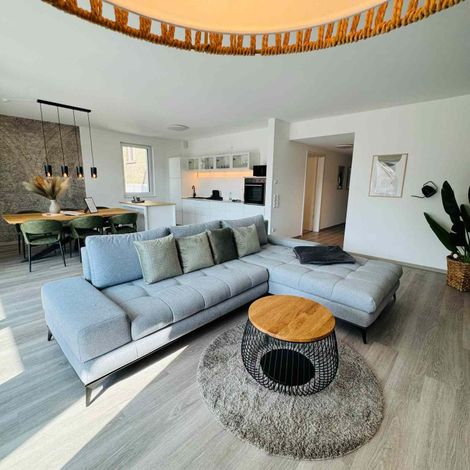 Helles, modernes Wohnzimmer mit grauer Eckcouch, rundem Couchtisch aus Holz, Essbereich und weißen Einbauschränken.