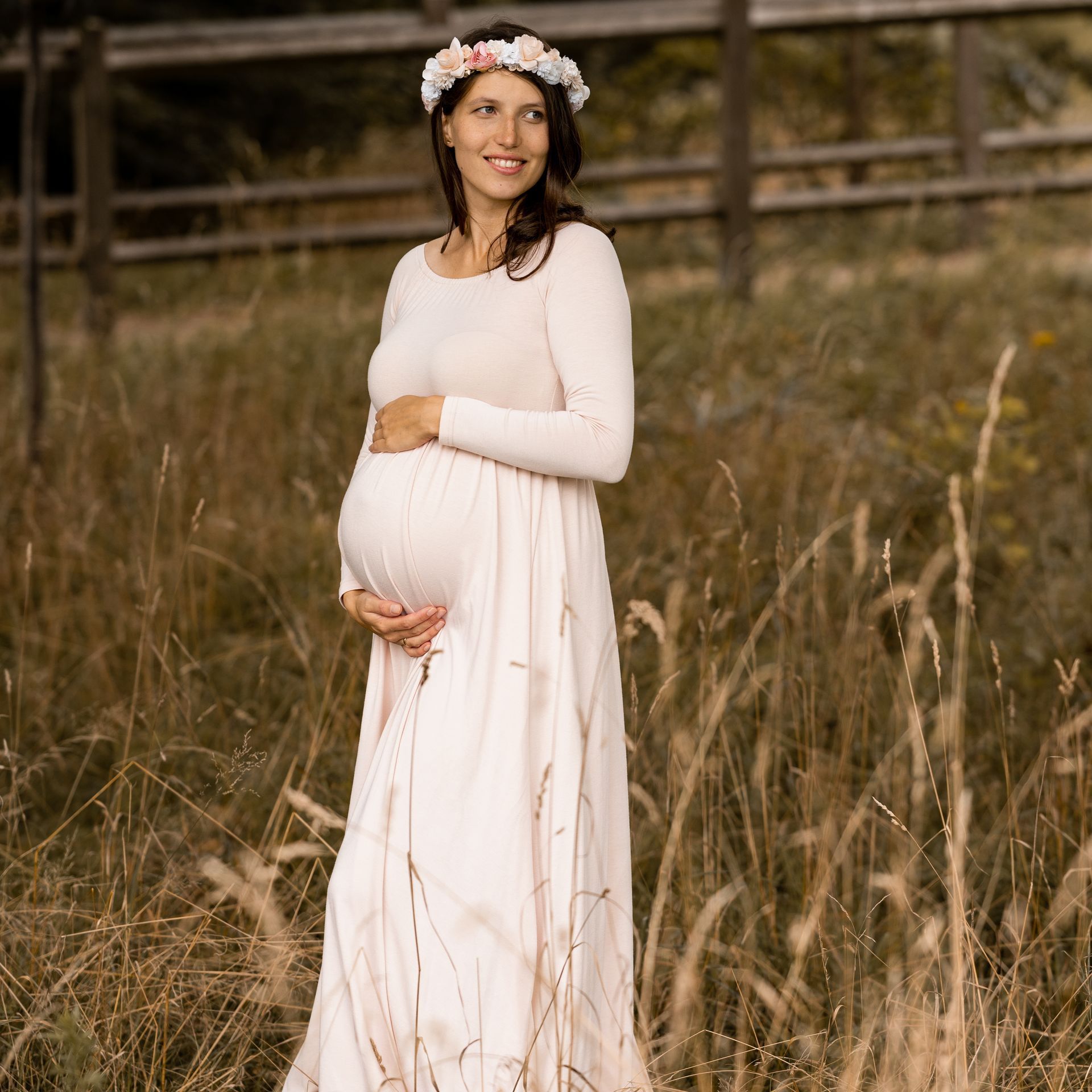 Eine schwangere Frau in einem rosa Kleid steht auf einem Feld und hält ihren Bauch.