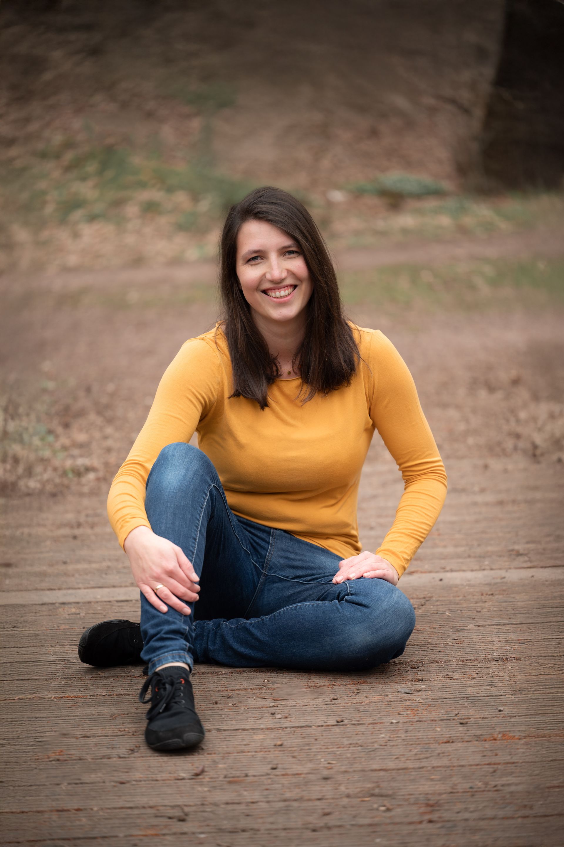 Marie in einem gelben Pullover sitzt mit überkreuzten Beinen auf dem Boden und lächelt.
