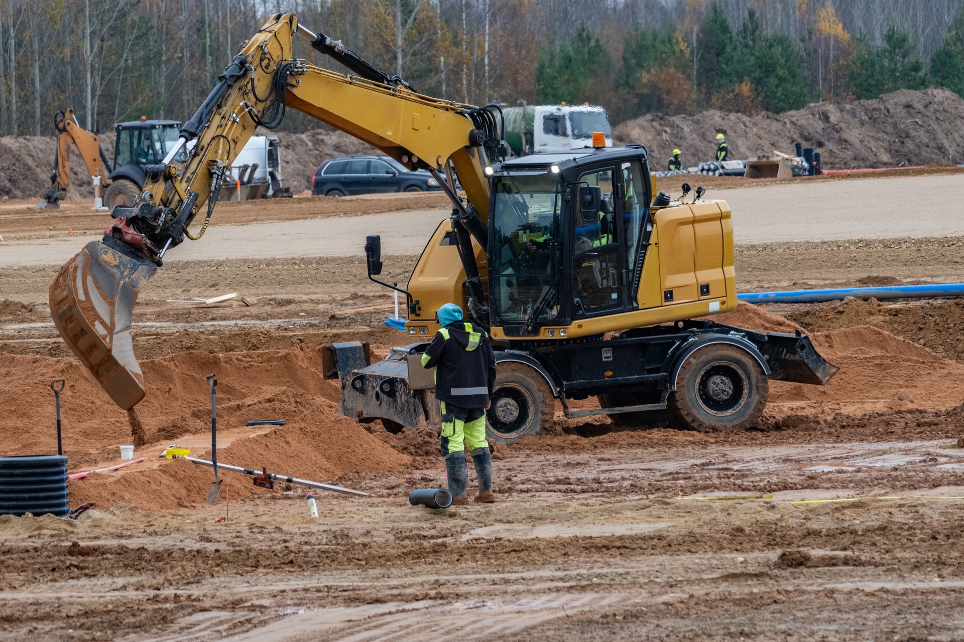 Une pelleteuse jaune creuse un terrain vague ; un ouvrier du bâtiment en tenue de sécurité se tient à proximité.