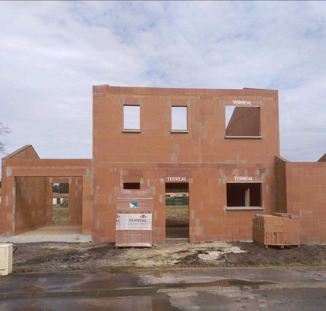 Construction d'un bâtiment en briques de deux étages, avec ouvertures pour fenêtres et portes. Un garage y est attenant. Ciel nuageux.