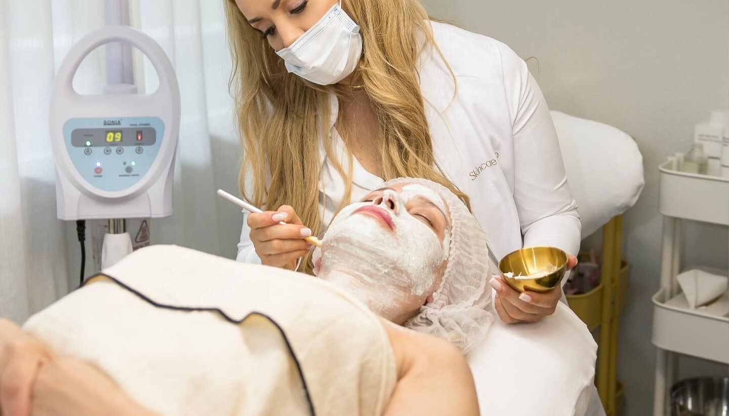 Un praticien en blouse blanche applique un masque facial à un client allongé sur une table de traitement dans un cadre clinique.