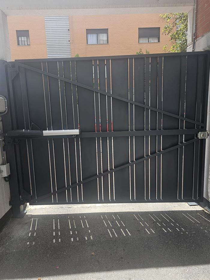 Una puerta negra está abierta a un camino de entrada con un edificio al fondo.