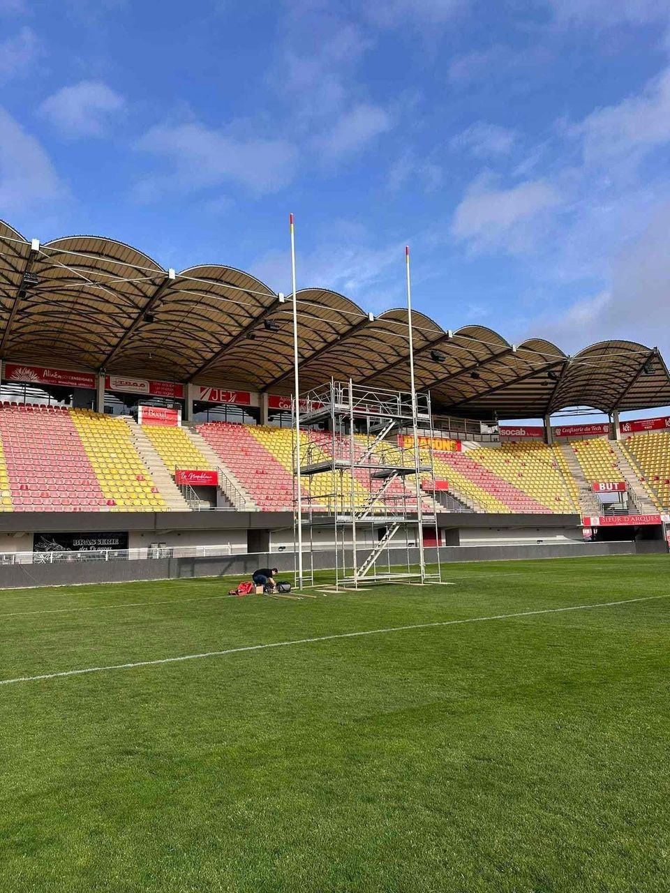 Stade de rugby vide, avec ses poteaux de but, un échaffaudage et ses sièges colorés sous un ciel bleu