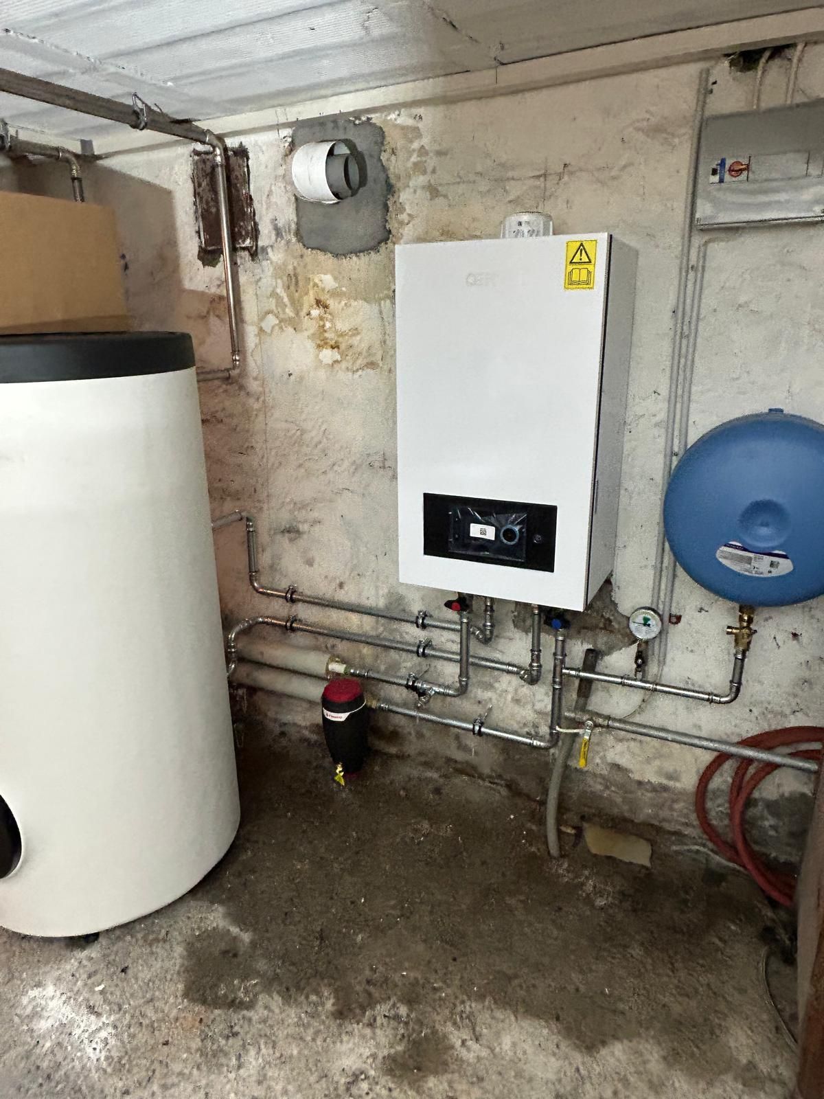 Une chaudière et un réservoir d'eau dans un sous-sol, avec des tuyaux, des composants électriques et un récipient sous pression.