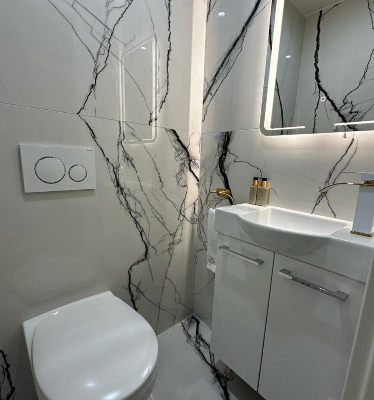 Petite salle de bains blanche avec carrelage mural imitation marbre, toilettes blanches, meuble-lavabo et robinetterie dorée.